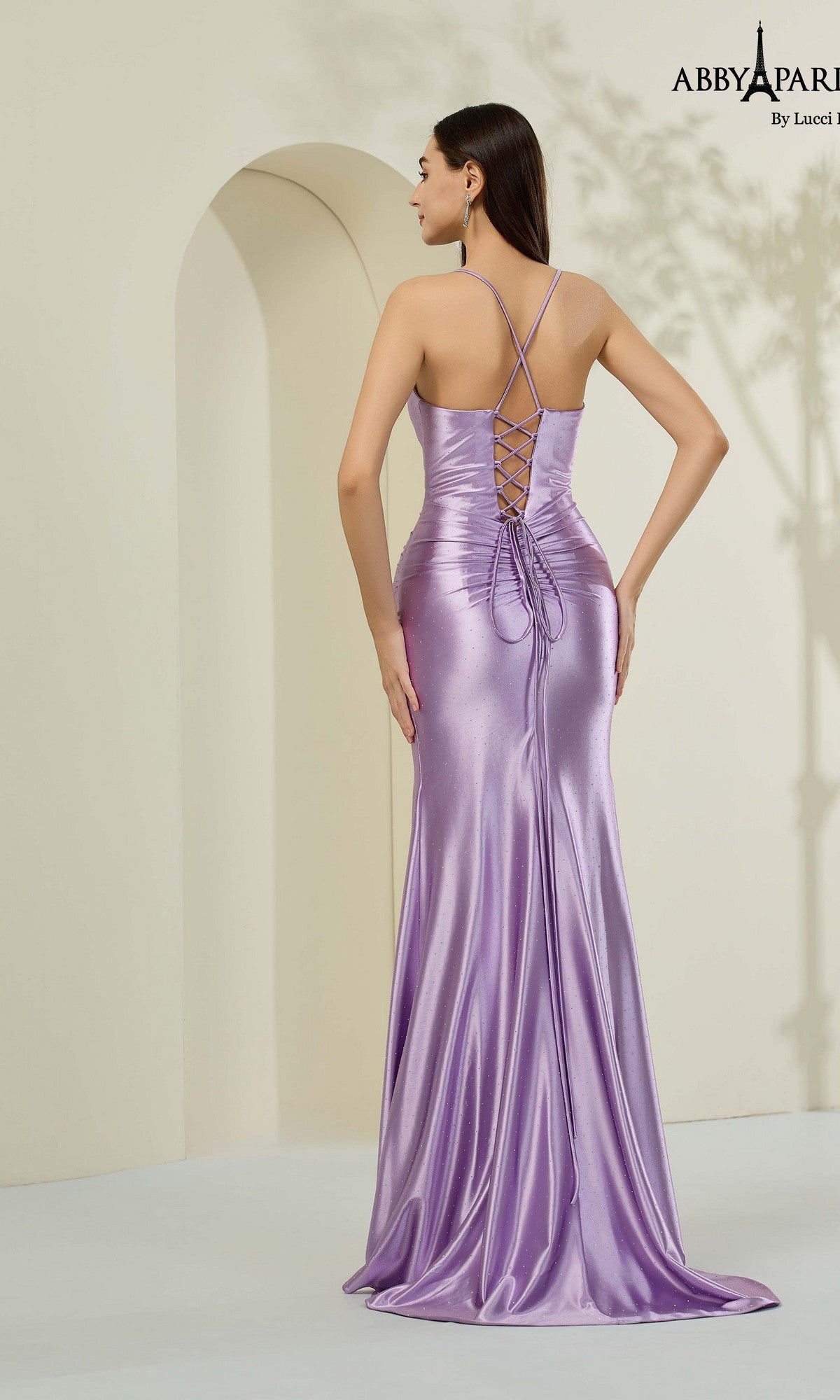 A woman stands facing away in a minimalist setting, wearing the Abby Paris 90375 long lavender satin prom dress featuring a shiny finish and lace-up back.